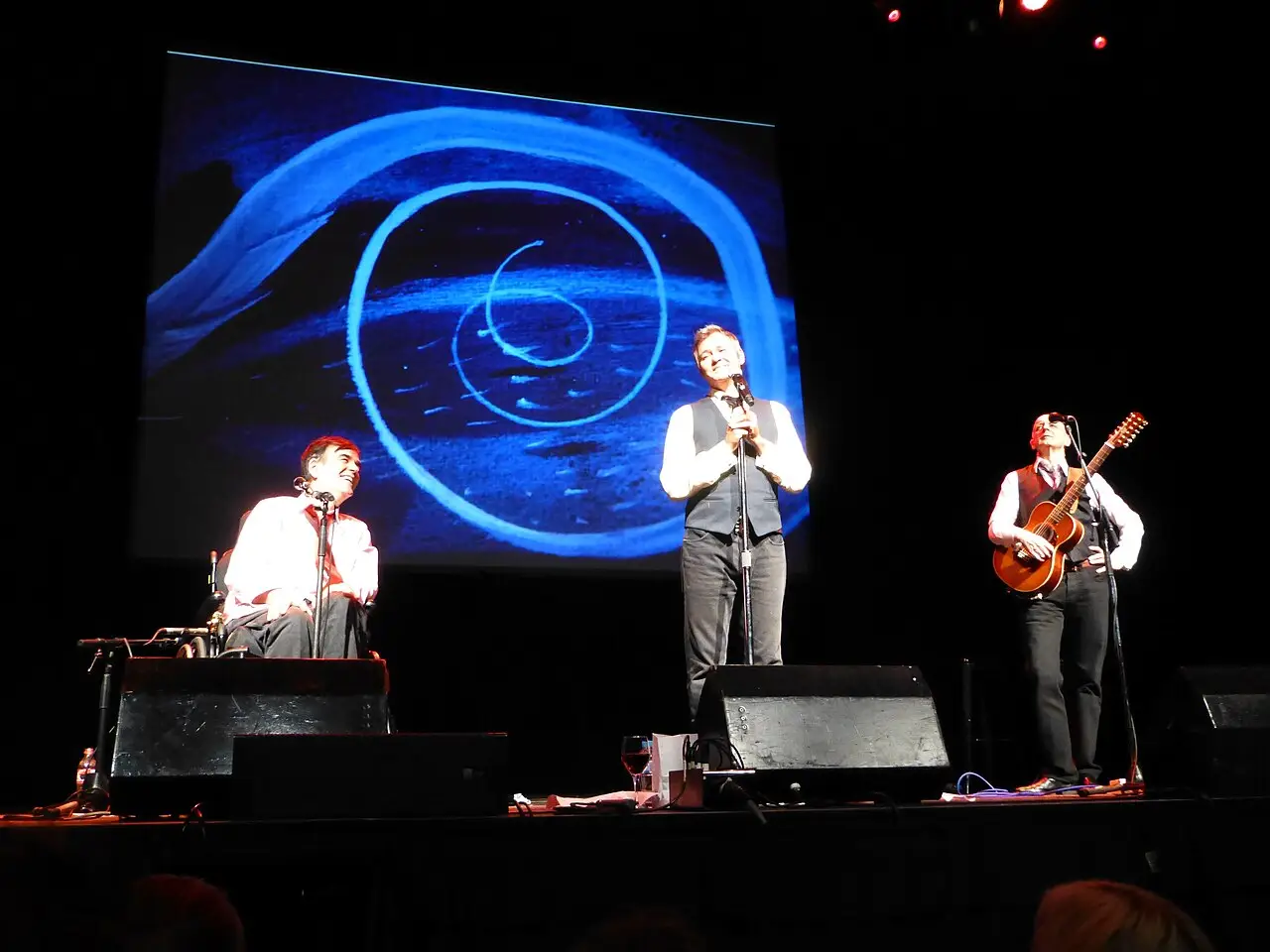 Three musicians play on a stage, one at a keyboard, one with a microphone, and one with a guitar, in front of a blue spiral screen.