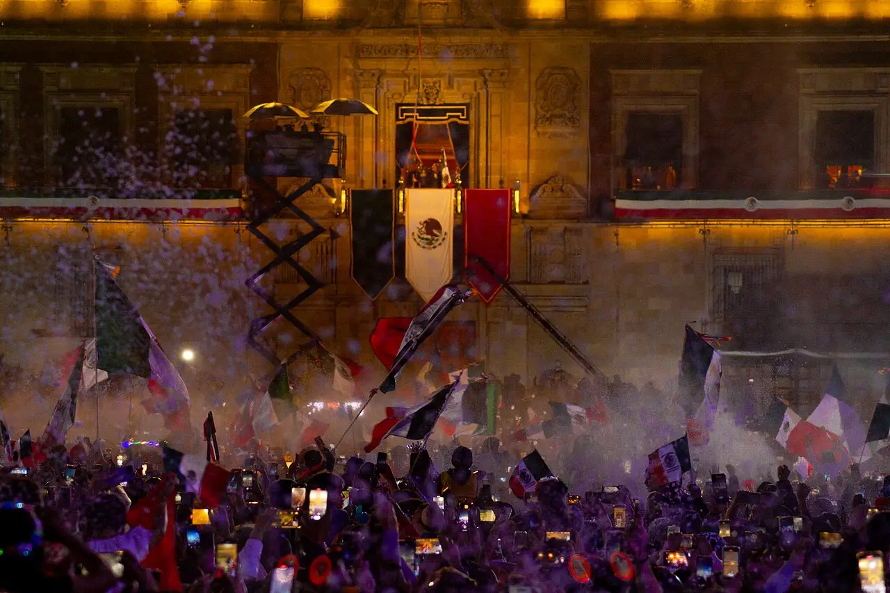 A multidão agita bandeiras diante do palácio, de cuja varanda pendem a bandeira do México e faixas verde e vermelha.