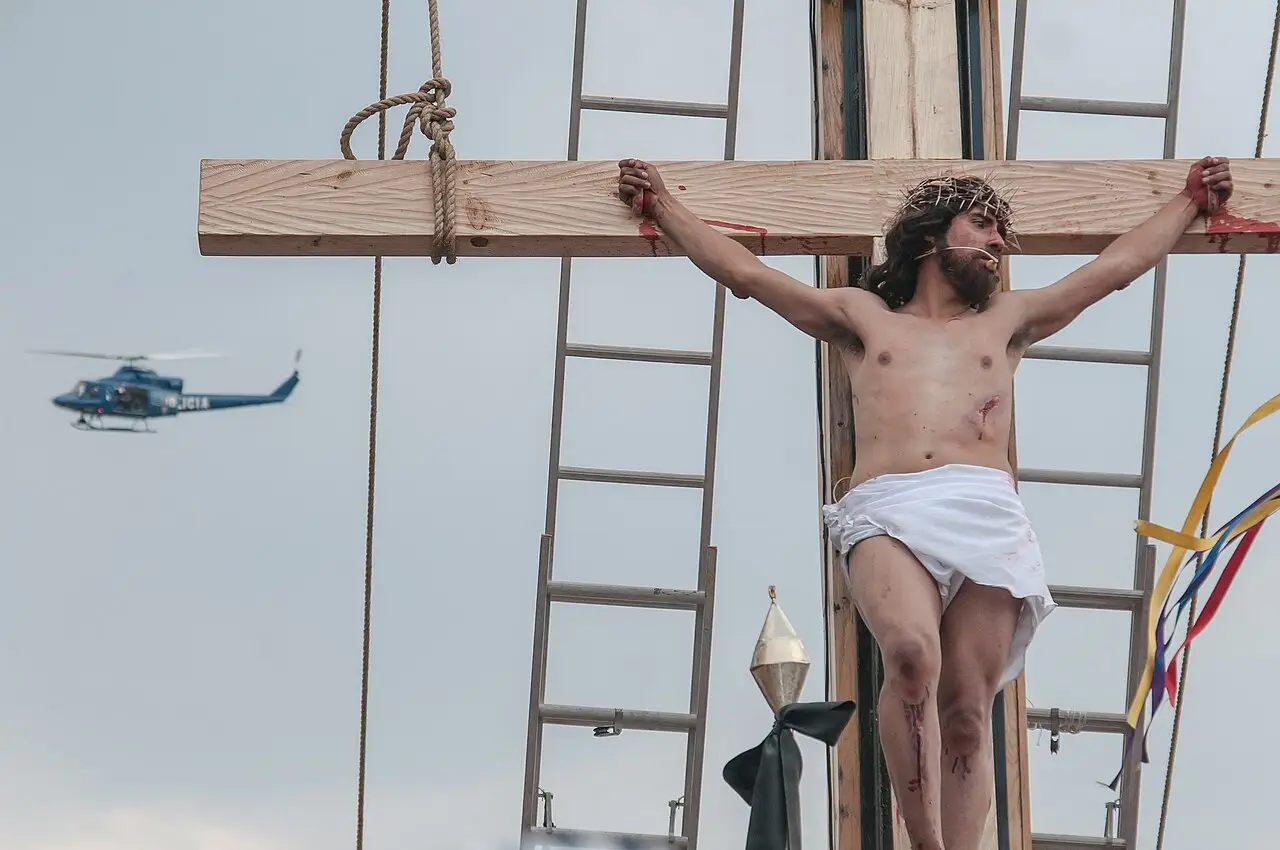 Um homem de pano branco está numa cruz de madeira, com uma escada ao lado e um helicóptero no céu.