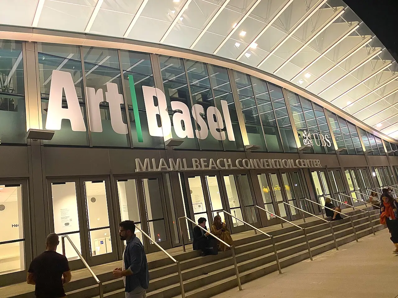 A fachada de vidro mostra as letras Art Basel, e pessoas estão na escada da entrada.
