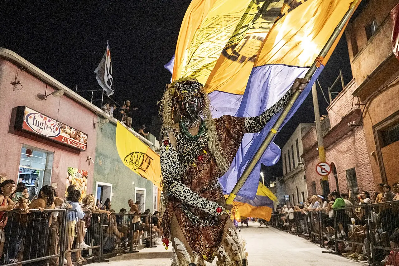 À noite, uma pessoa de fantasia leva uma bandeira amarela e azul no meio da rua, com o público atrás das grades.