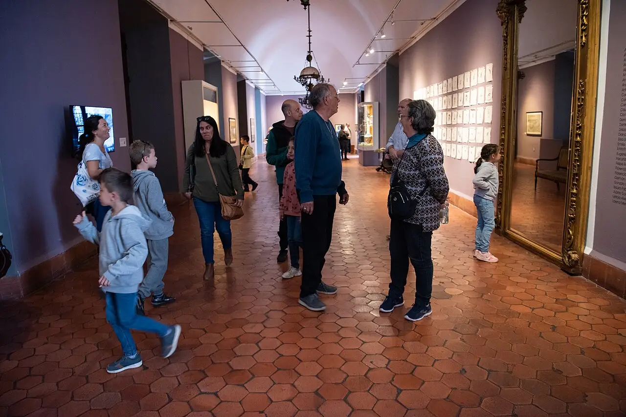 Visitantes percorrem um corredor do Cabildo, de chão hexagonal e paredes roxas, com quadros à direita.