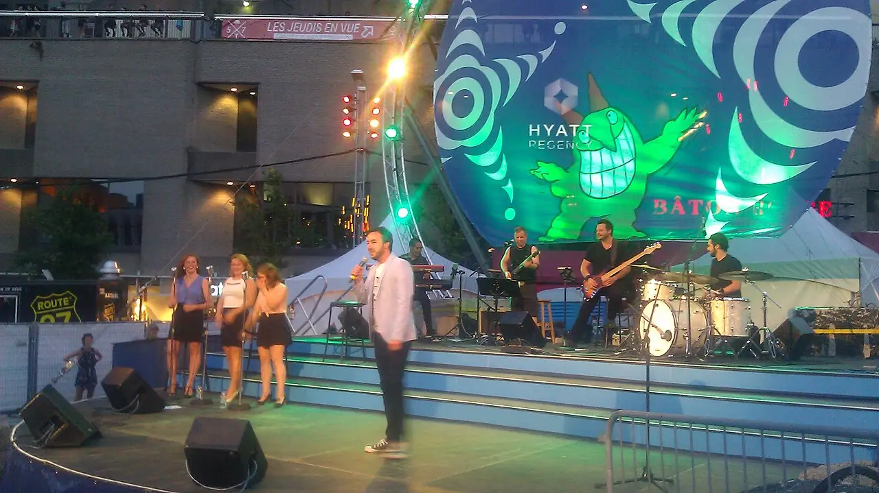 Num palco ao ar livre, um homem de camisa branca fala ao microfone, com a banda atrás e um painel verde e azul.