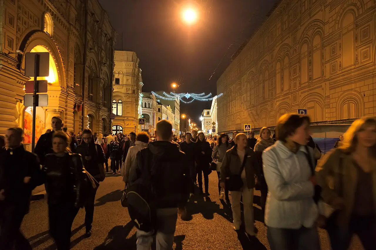 La nuit, une foule marche dans une rue entre des immeubles anciens, sous un lampadaire et des fils de lumière.