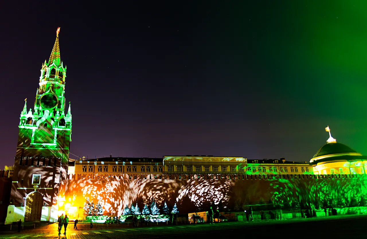 Le mur du Kremlin et la tour de l’horloge portent une projection verte, avec des sapins de lumière à la base et des silhouettes sur la place.