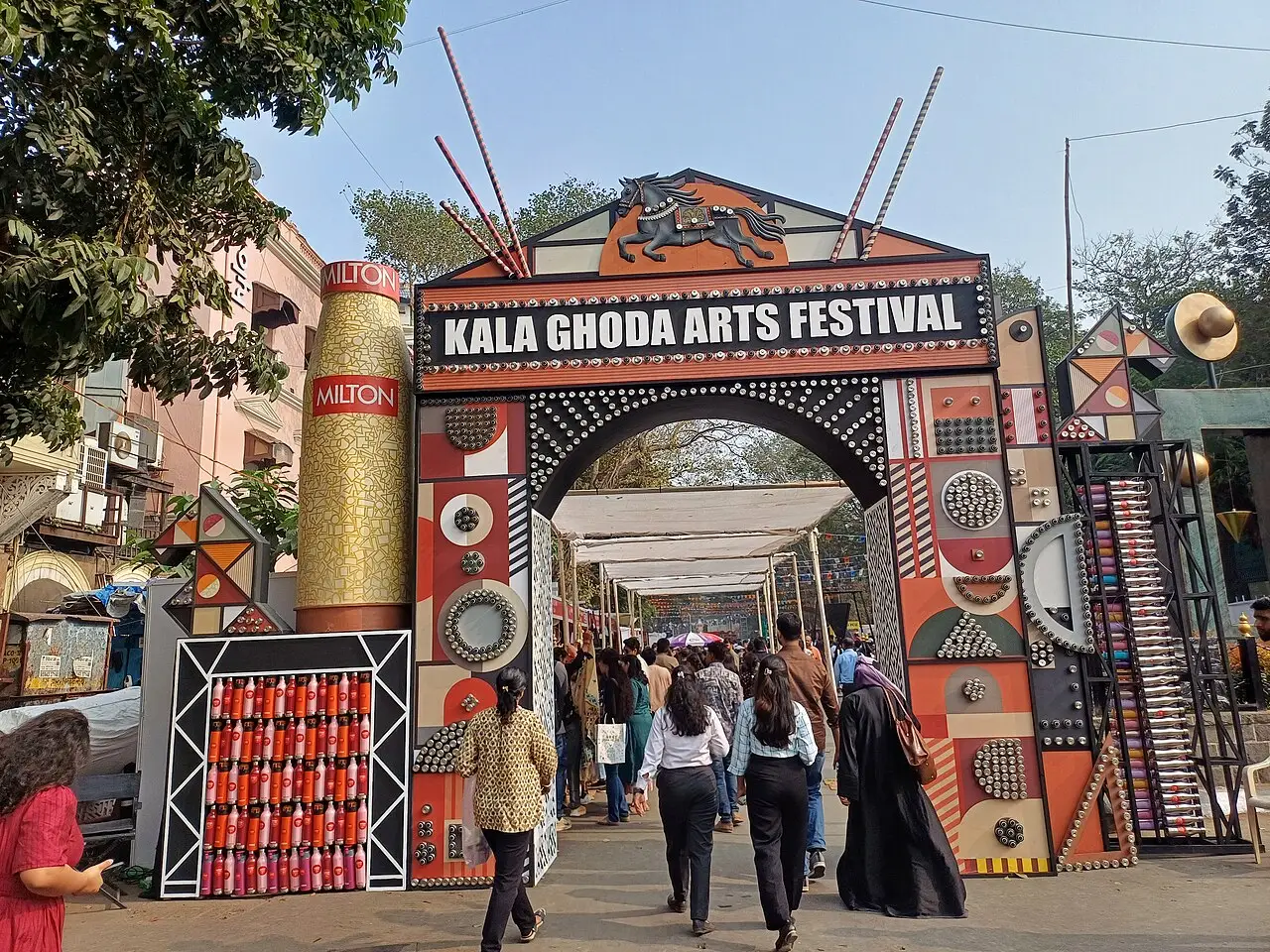 Pessoas passam sob um arco pintado com a faixa Kala Ghoda Arts Festival e desenhos em vermelho, preto e dourado.