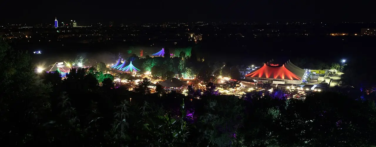 À noite, tendas iluminadas ocupam o parque, uma delas vermelha, com luzes verdes e roxas e a cidade no horizonte.