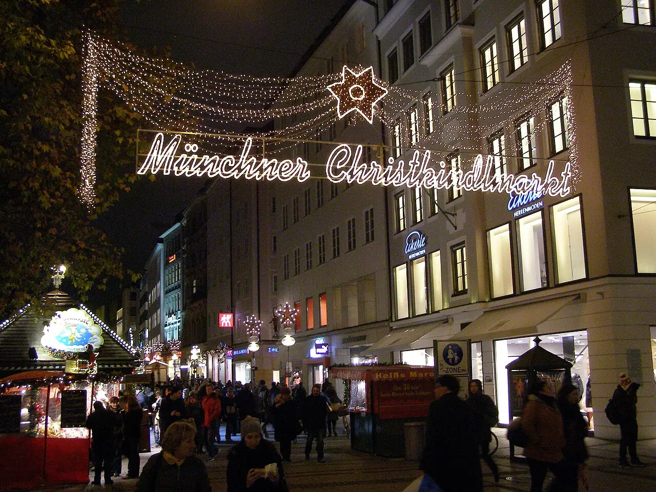 À noite, pessoas caminham sob o letreiro luminoso Münchner Christkindlmarkt, com estrelas de luz e uma barraca à esquerda.