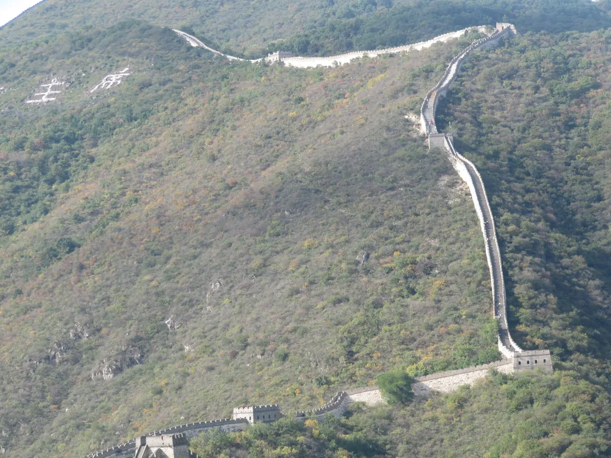 A Muralha da China serpenteia pelas montanhas de Badaling, com torres e visitantes pequenos sobre as escadas.