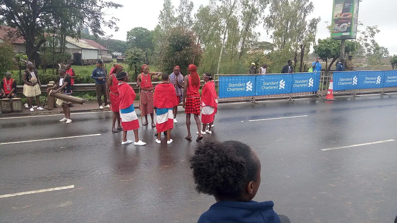 Pessoas de manto vermelho estão na pista molhada, com barreiras azuis atrás e uma criança no primeiro plano.