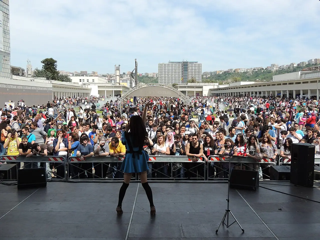 De costas, uma pessoa de saia azul está no palco ao ar livre, diante de uma plateia grande, com prédios e morros ao fundo.