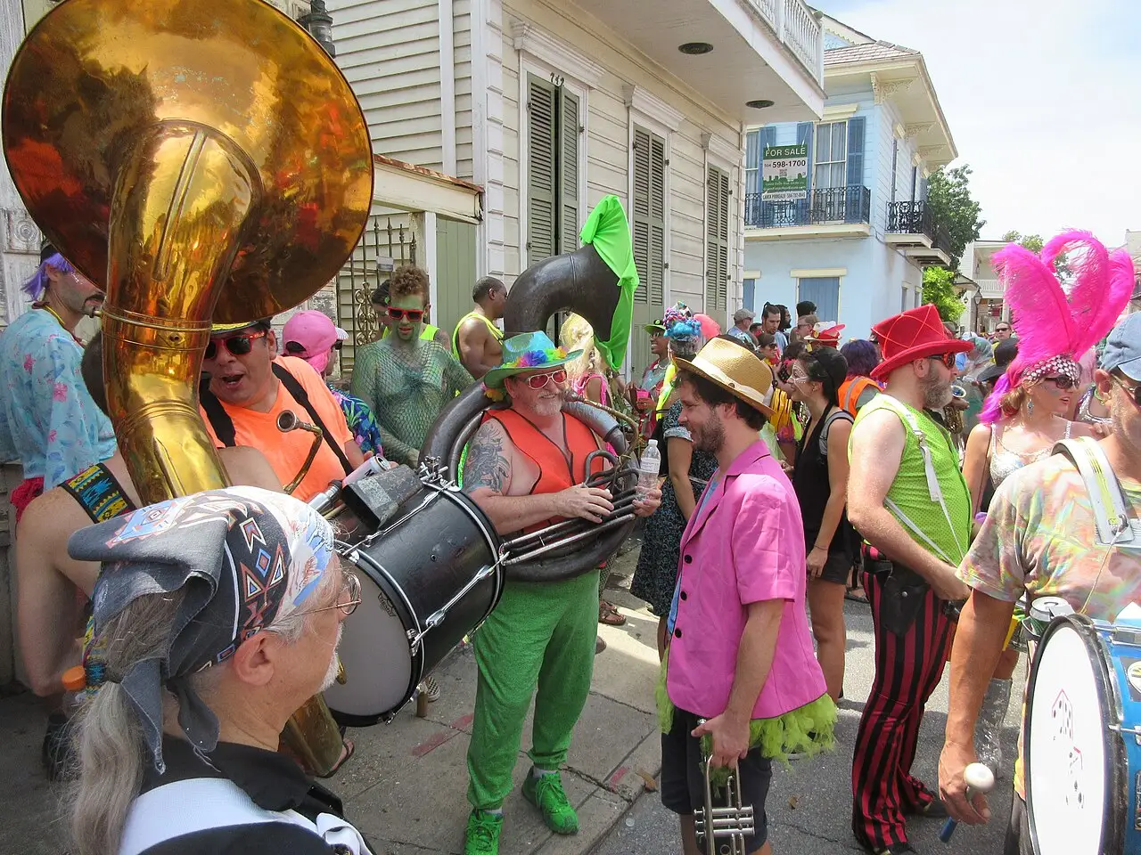 Músicos de roupa colorida tocam metais e tambor na rua, com um sousafone dourado, diante de uma casa clara.