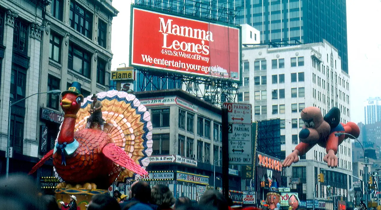Um balão gigante de peru e outro balão passam entre prédios altos, sob o letreiro vermelho da Mamma Leone's.