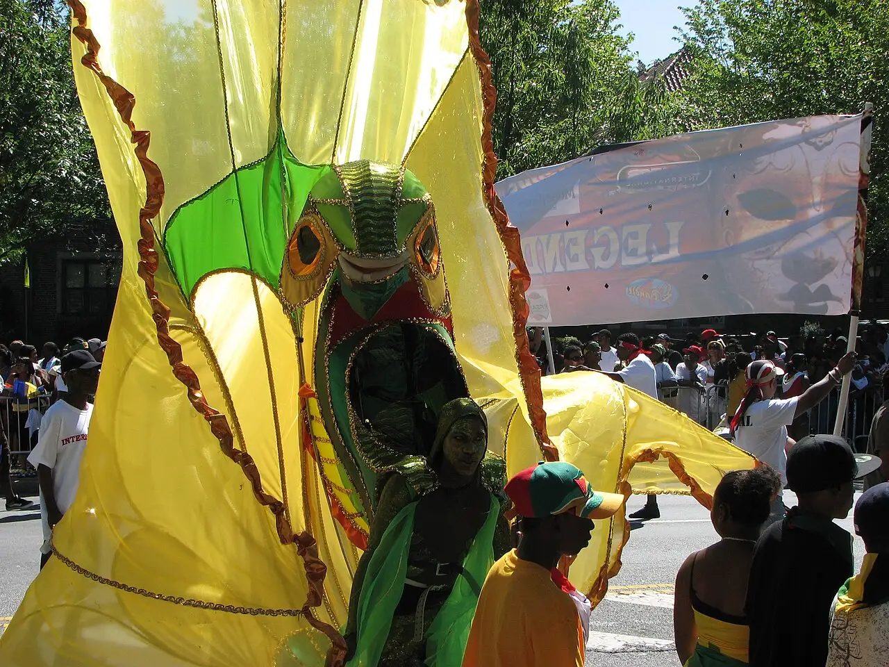 Pessoas num grande traje amarelo e verde, com uma máscara verde, caminham na rua, com público atrás.