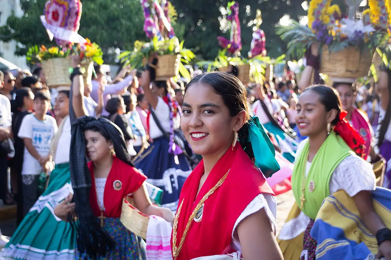 Mulheres de vestido bordado e blusa vermelha caminham num desfile, com cestos de flores acima da multidão.