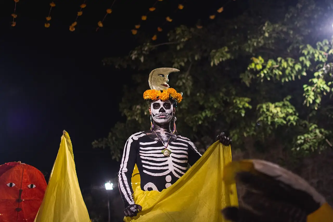 À noite, uma figura grande de esqueleto, com chapéu de flores laranjas, segura um pano amarelo, sob árvores com luzes.