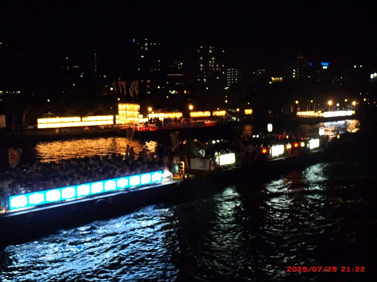 À noite, barcos com luzes azuis e lanternas levam gente num rio, com os prédios da cidade ao fundo.