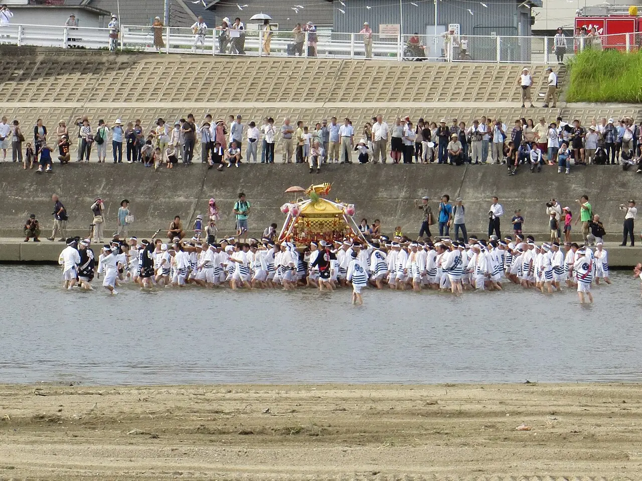 Pessoas de branco carregam um santuário dourado portátil pela água do rio, e a multidão assiste da margem.