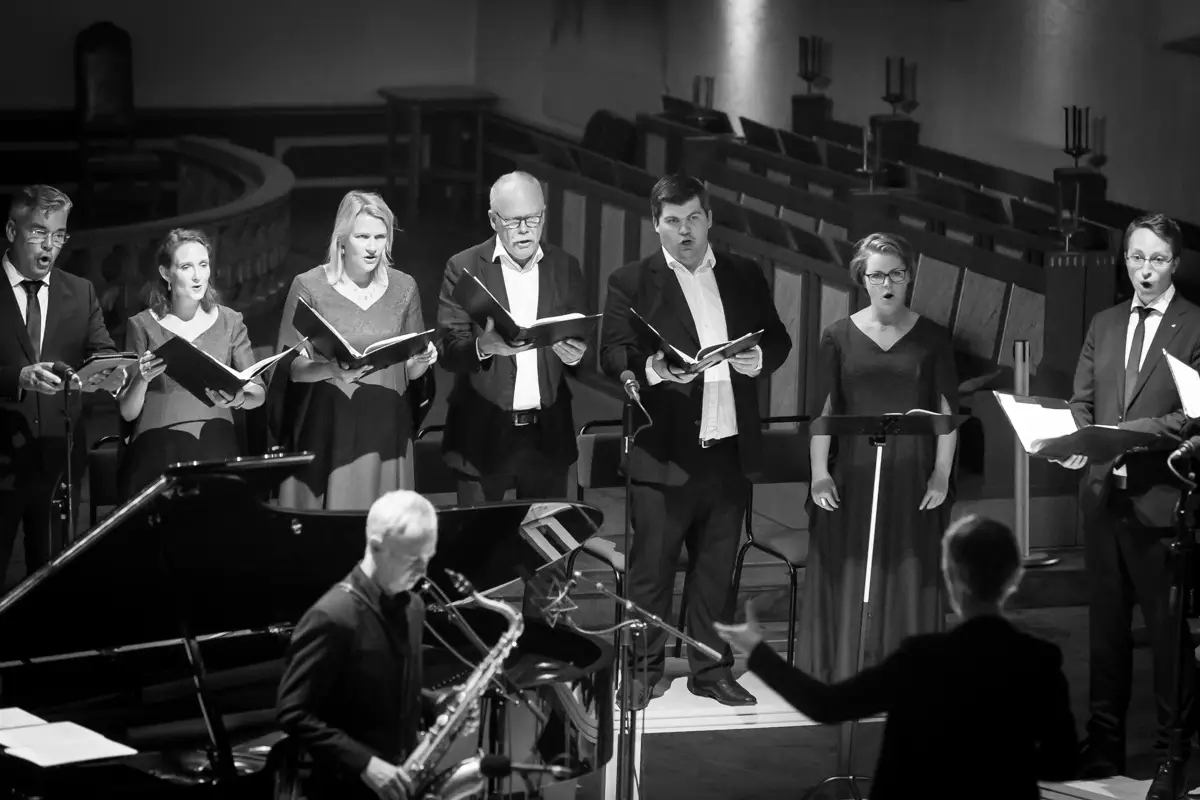 Une chorale, partition en main, chante, avec un saxophoniste et un chef devant, sur une photo en noir et blanc.