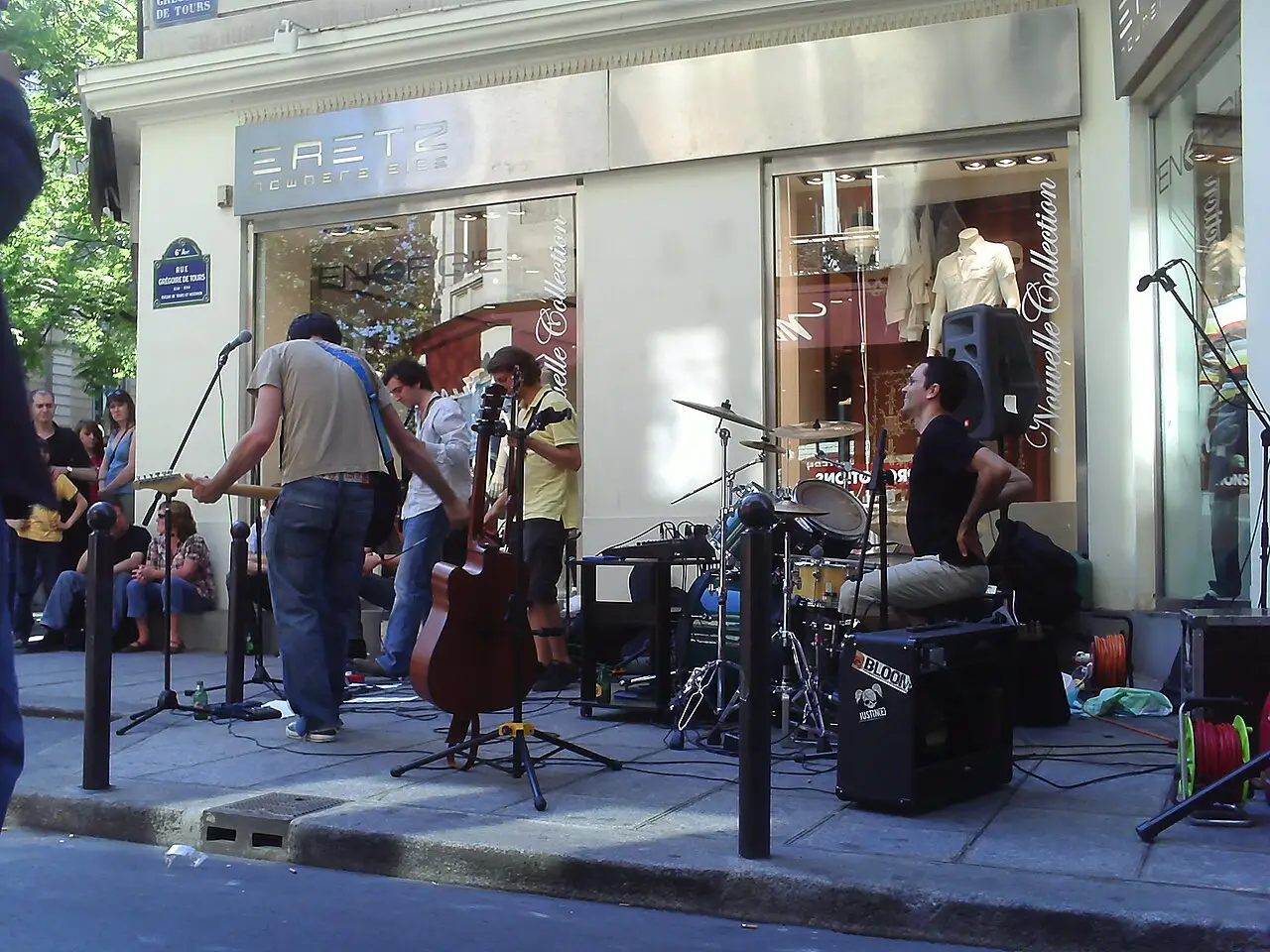 Uma banda com guitarra, contrabaixo e bateria toca na calçada diante de uma vitrine.
