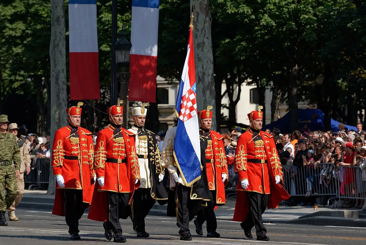 Soldados de casaco vermelho e cinto branco marcham com bandeiras, entre elas as da França, diante do público.