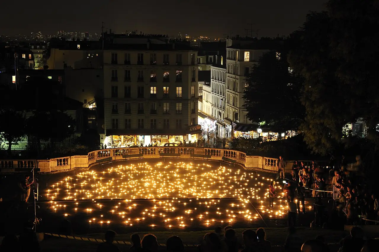 De noche, muchas velas cubren el suelo de una plaza, con gente en el borde y ventanas encendidas detrás.