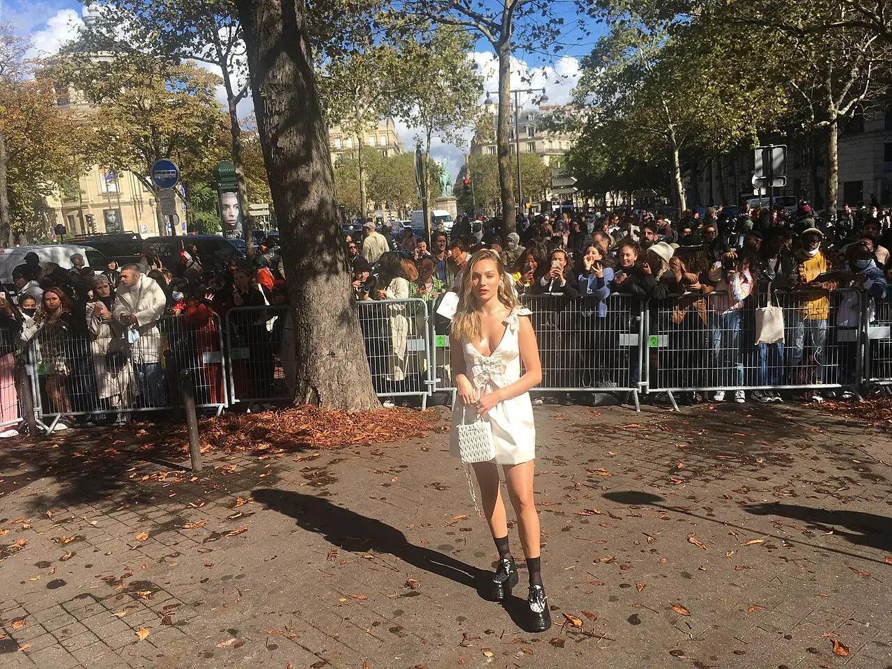 Uma mulher de vestido branco curto está na rua diante de uma multidão atrás de grades, com árvores ao redor.