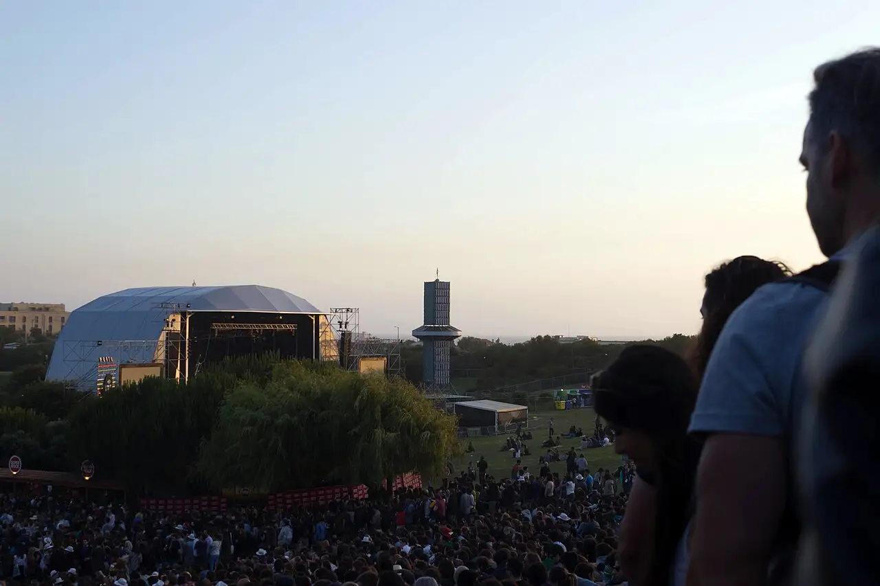 O público, visto de costas, olha um palco ao ar livre, com uma cúpula prateada e uma torre ao fundo, ao anoitecer.