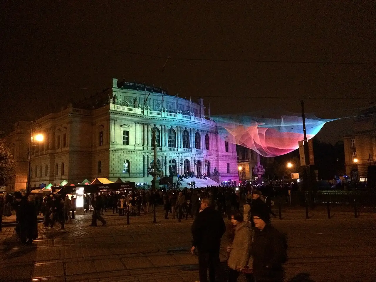 O Rudolfinum está iluminado de verde e de rosa, com uma teia de luz sobre a praça e pessoas em volta.
