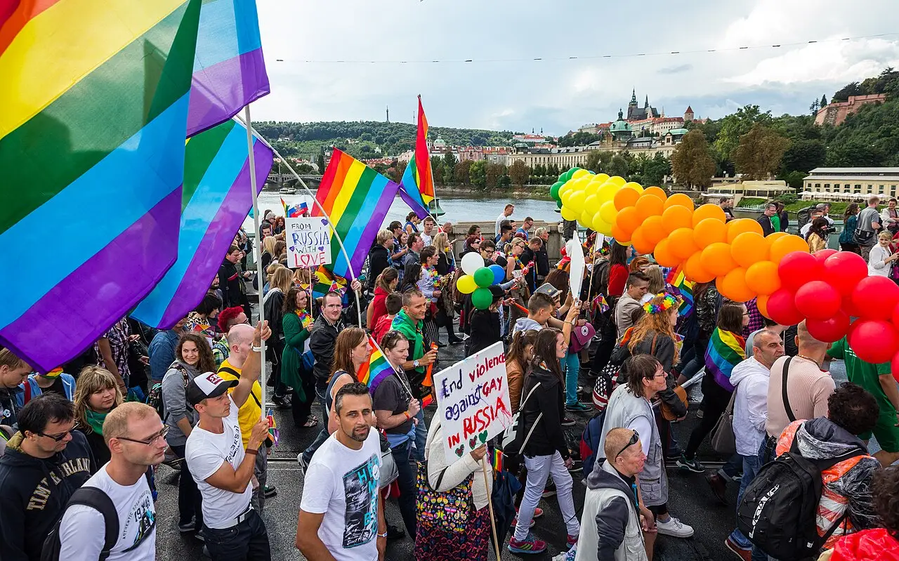The parade crosses the bridge with rainbow flags and a row of orange, yellow and red balloons, and the river is behind.
