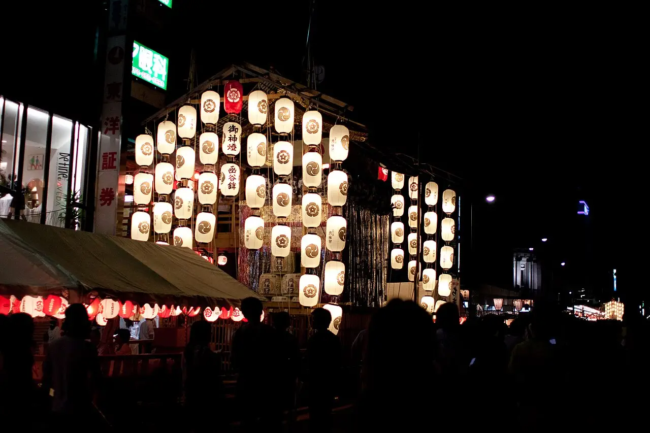 Um carro do festival coberto de lanternas brancas de papel está na rua à noite, com pessoas ao redor.