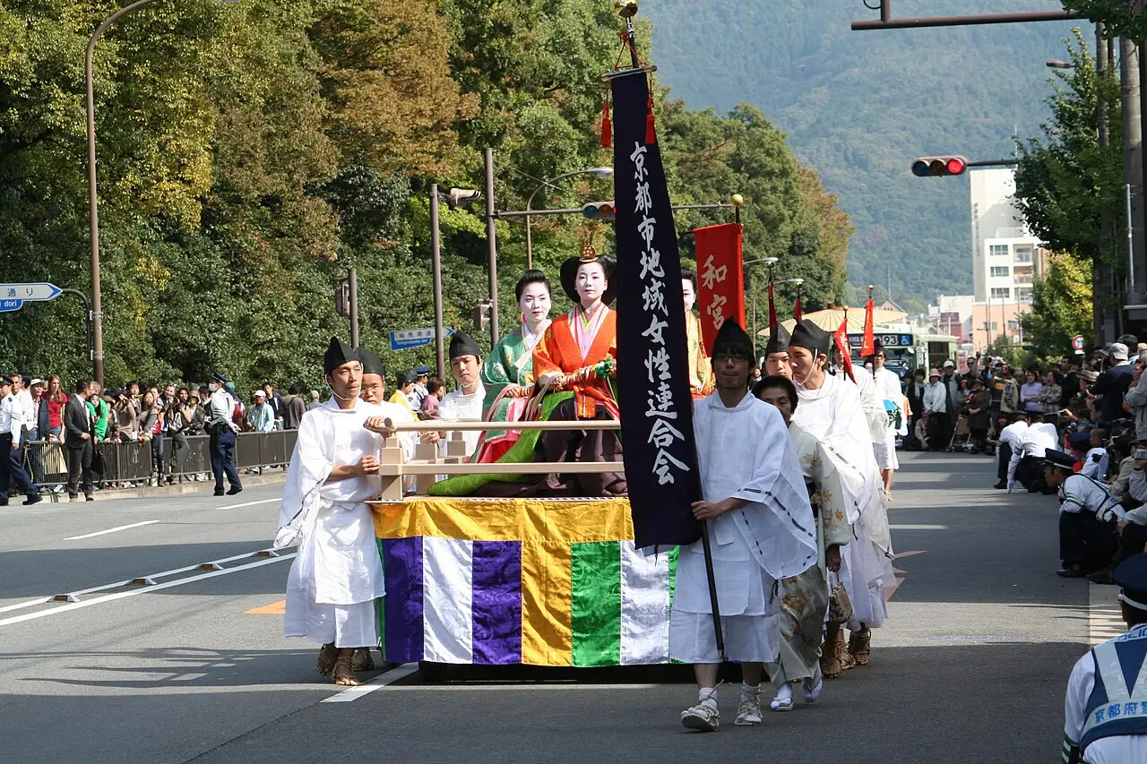 Um carro do desfile leva pessoas de traje histórico, com acompanhantes de branco e estandartes, por uma avenida.