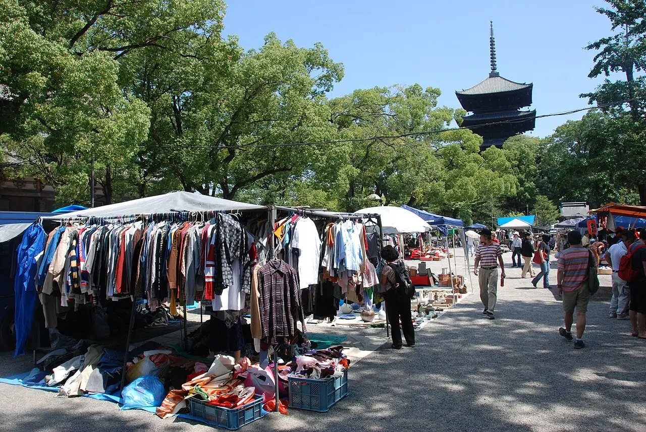 Barracas de roupa ocupam o pátio, a pagoda fica ao fundo e pessoas passam entre as mesas.