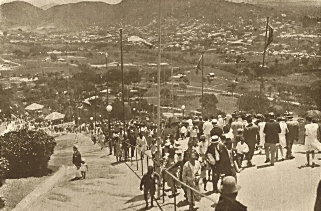 Fotografia em sépia da escadaria da Penha, com pessoas subindo e a paisagem do Rio de Janeiro ao fundo, em 1928.