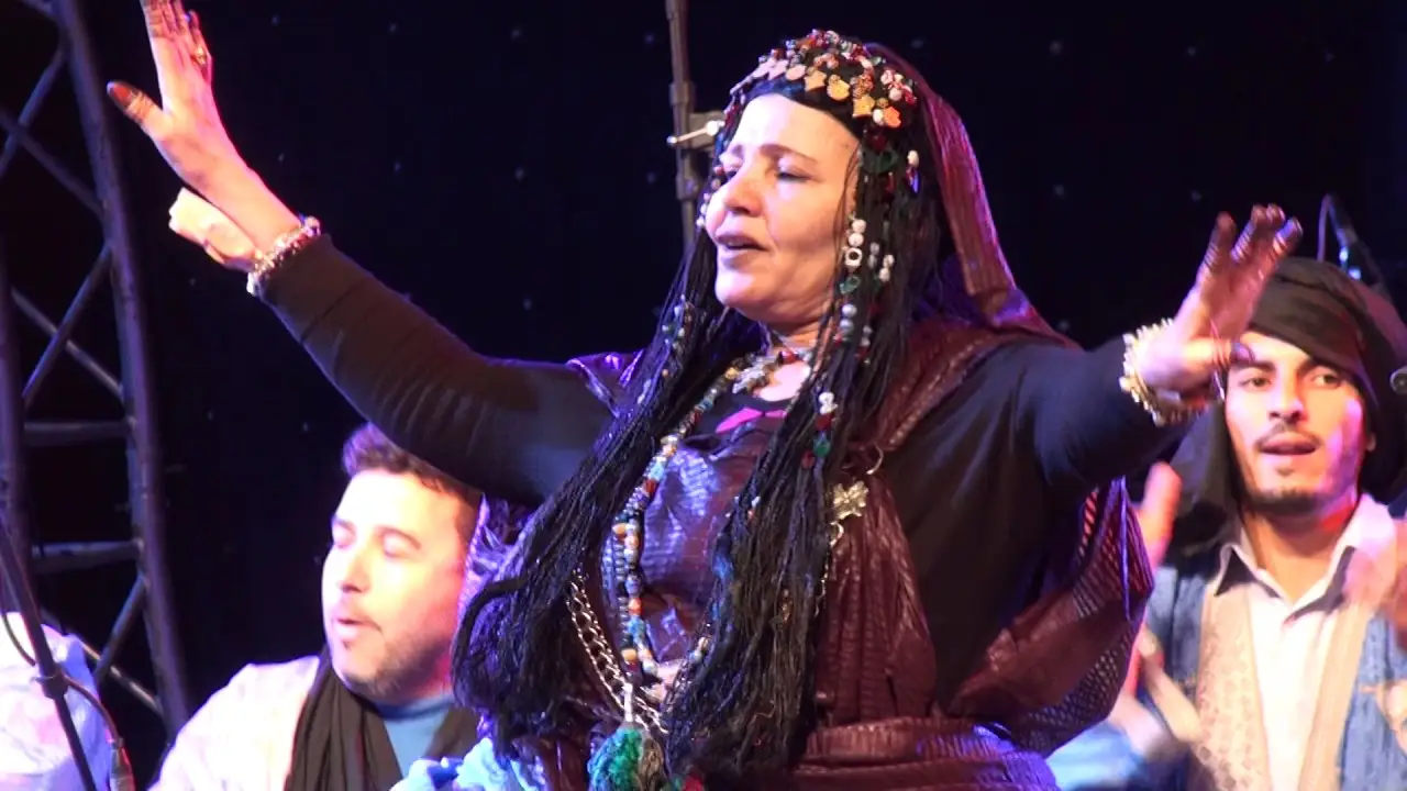 Uma dançarina de guedra está no palco de braços erguidos, com dois músicos ao lado.