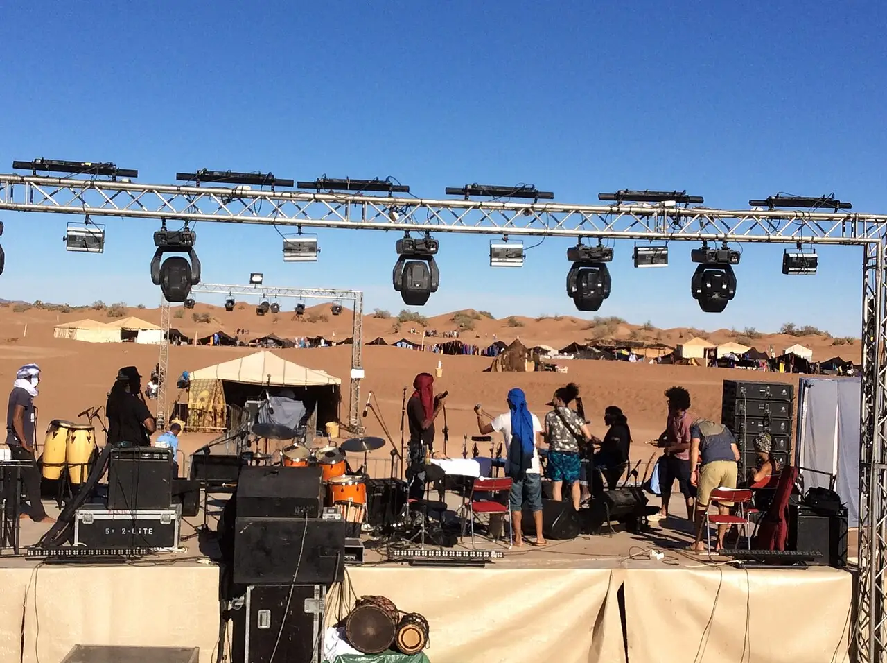 Músicos ocupan un escenario en el desierto, bajo una estructura de focos, con tiendas al fondo.