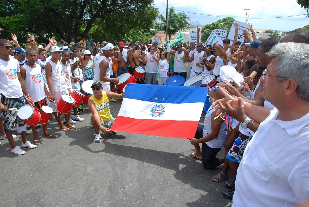 A multidão de branco ocupa a rua, com tambores vermelhos e uma bandeira aberta, de brasão, na frente.