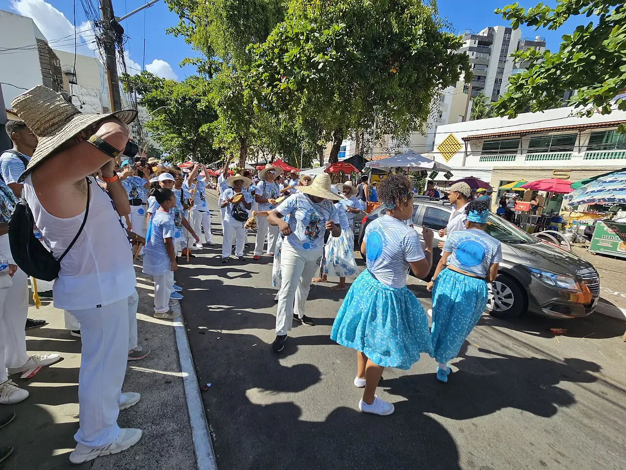 Mulheres de saia azul caminham na rua com o cortejo de branco, e carros ficam estacionados junto ao meio-fio.