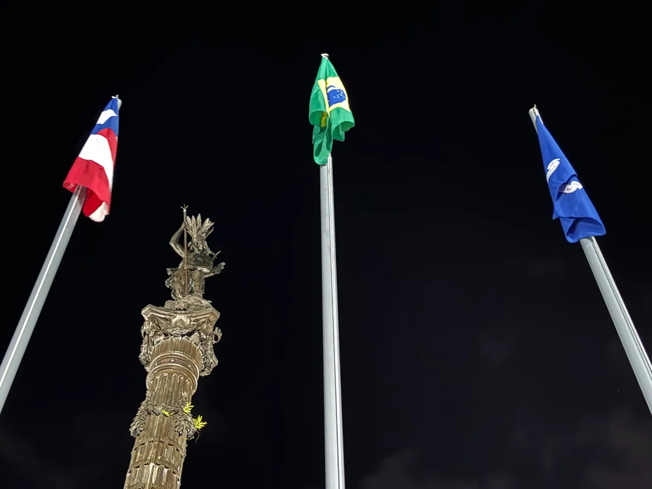 O monumento ao Caboclo no Campo Grande, em Salvador, na noite de 2 de julho de 2024, entre as bandeiras da Bahia, do Brasil e da cidade.