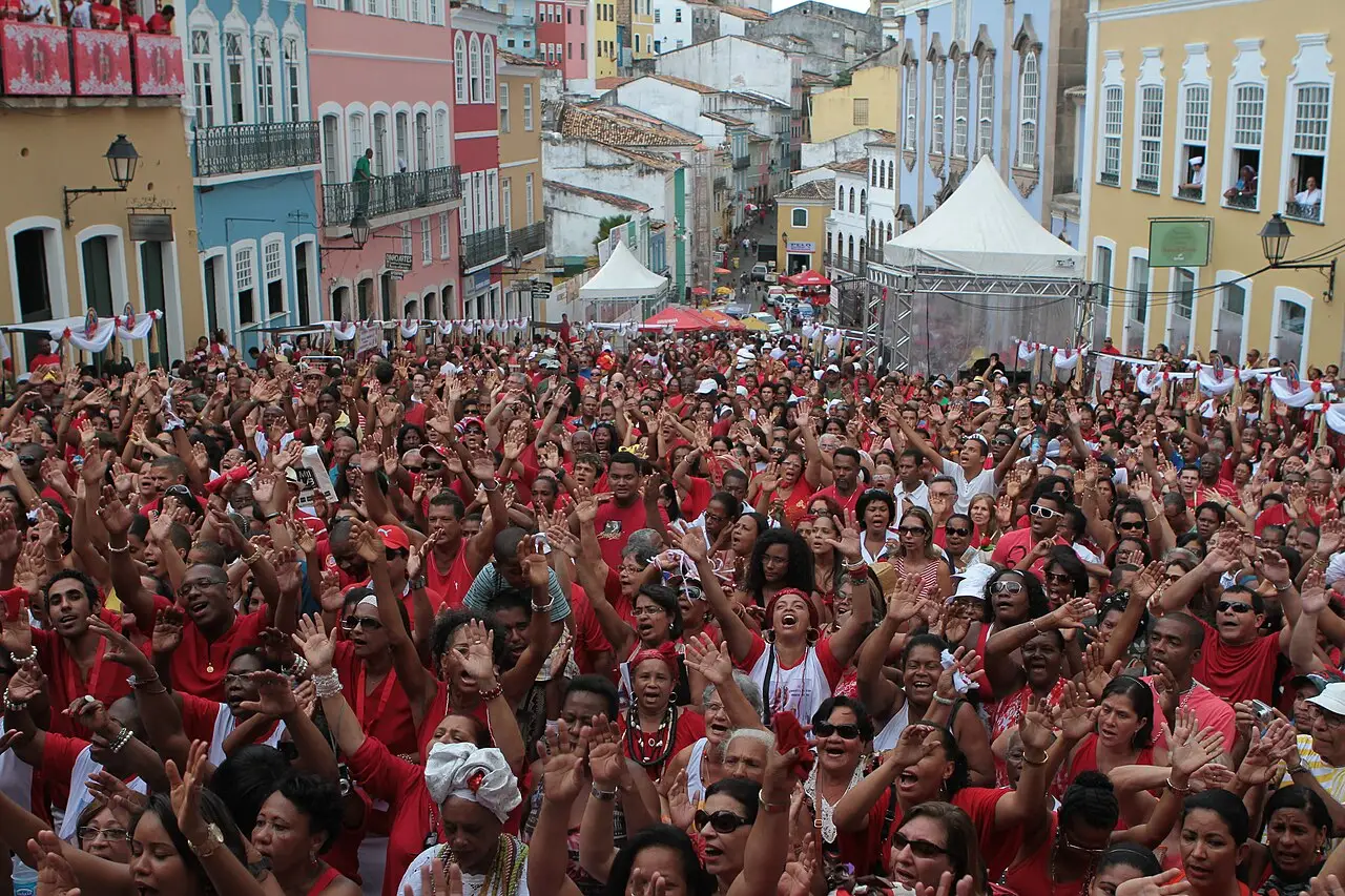 A multidão de vermelho enche a rua entre casas coloridas, com as mãos erguidas e uma tenda branca mais adiante.
