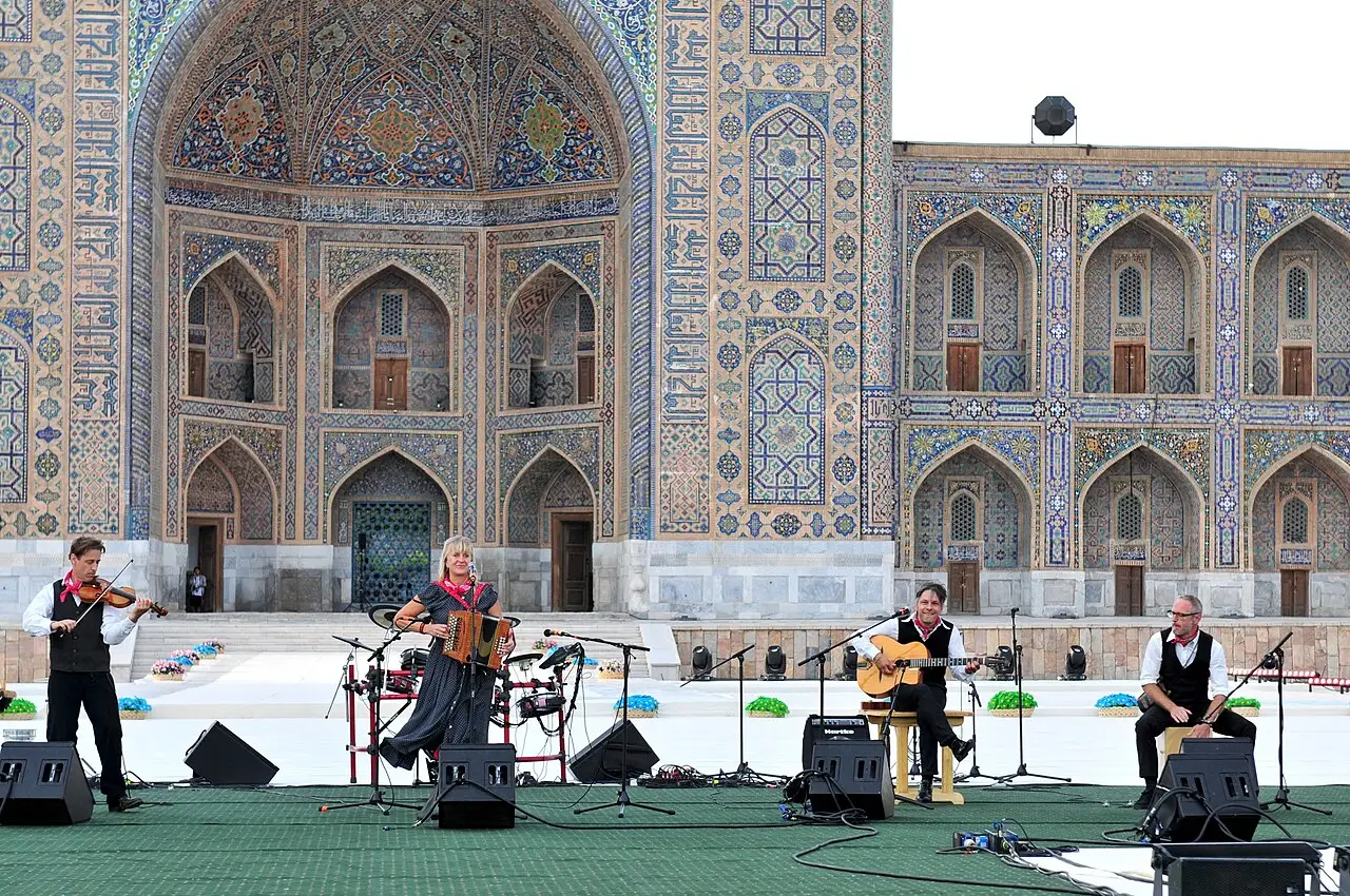 Quatro músicos tocam num palco ao ar livre diante de um arco revestido de azulejos azuis e dourados.
