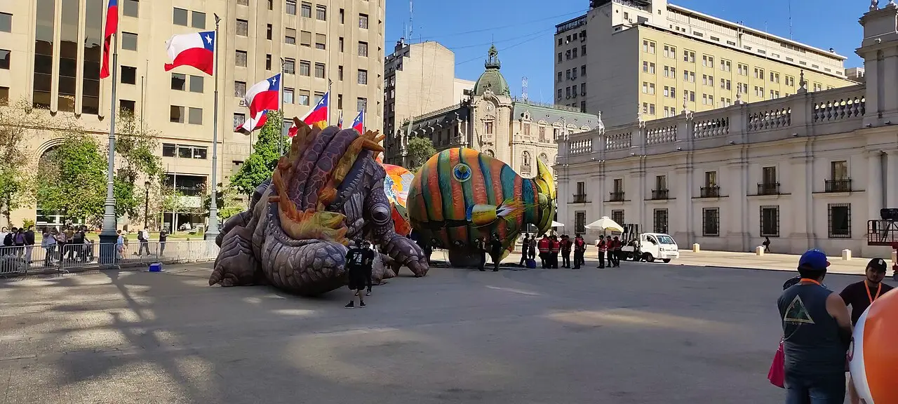 Duas figuras gigantes, uma de escamas e outra redonda verde e amarela, estão na praça, com bandeiras do Chile e prédios de pedra em volta.