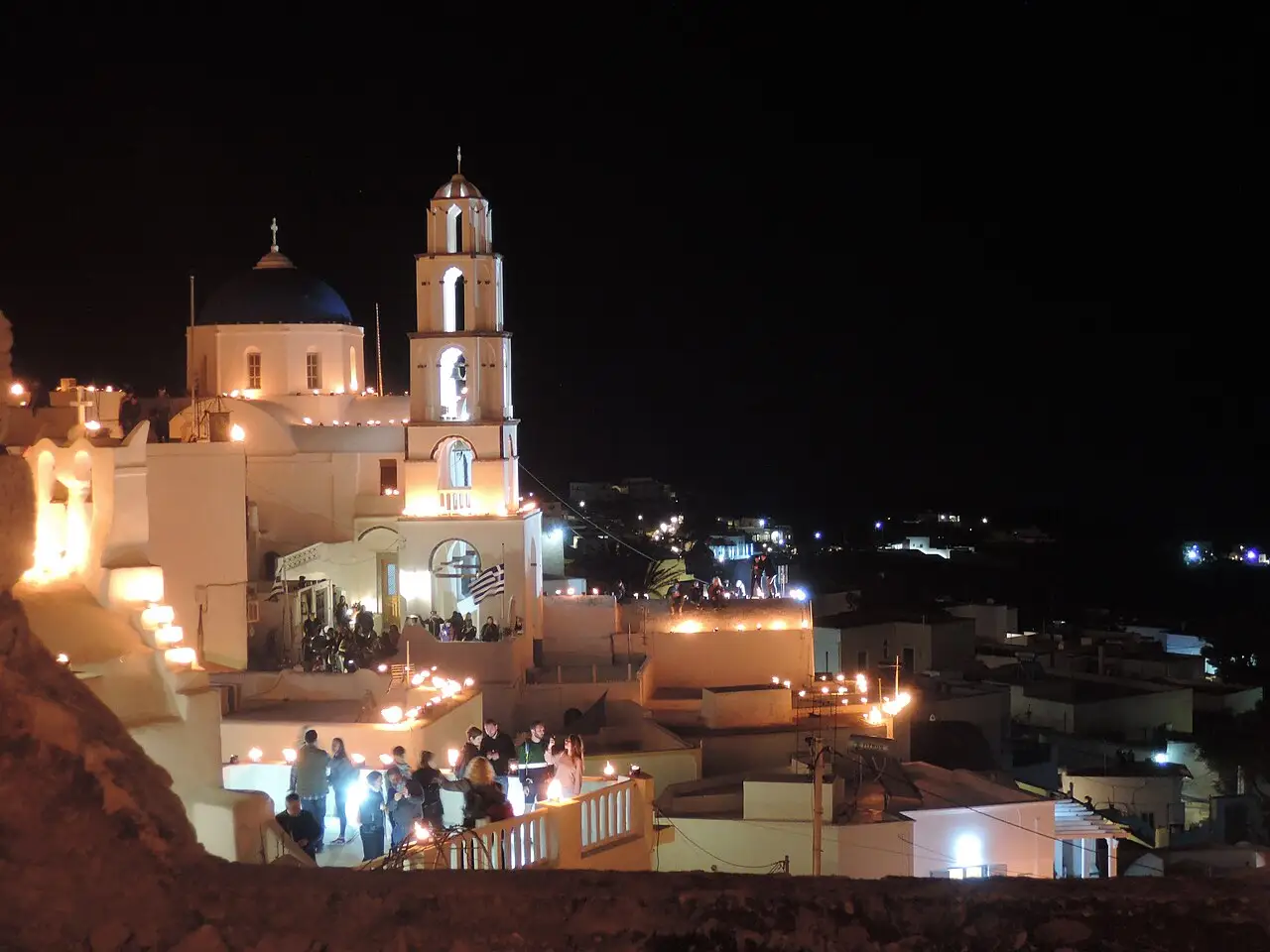 À noite, pessoas se reúnem nos terraços abaixo de uma igreja branca de cúpula azul e campanário iluminado.