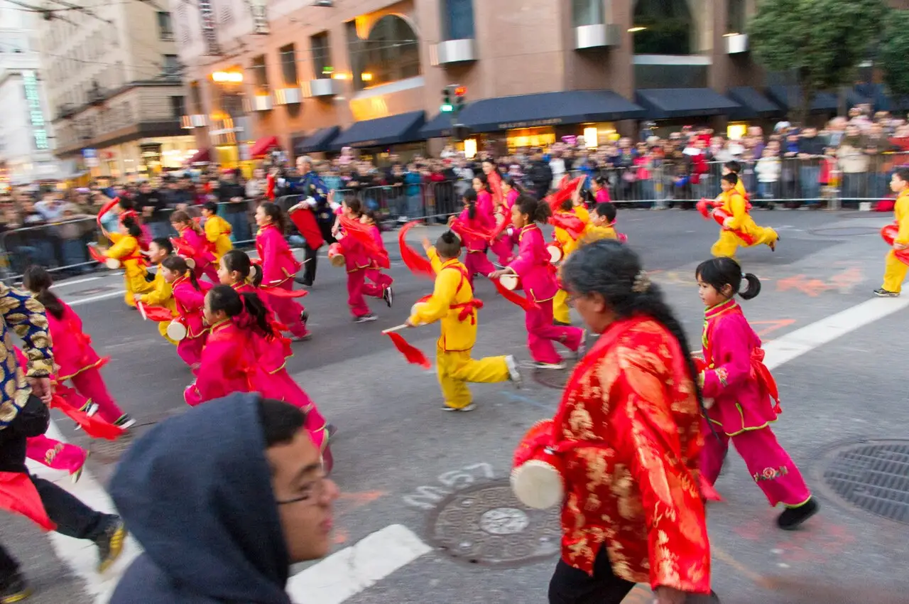 Dançarinos de rosa e amarelo atravessam a rua, uma mulher de vermelho e dourado vem na frente, e o público olha atrás das grades.