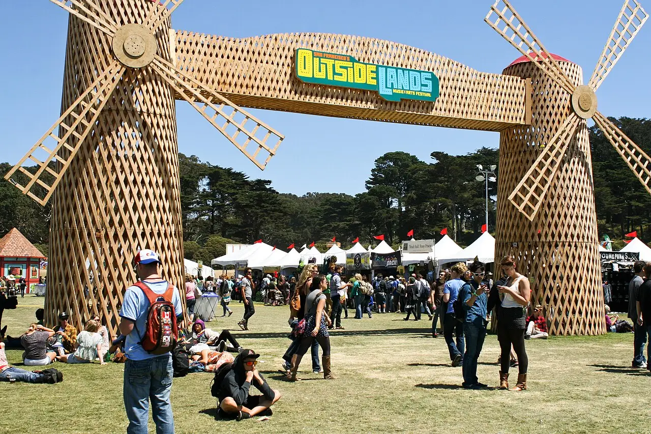 Dois moinhos de madeira sustentam um arco com o letreiro Outside Lands, e pessoas e barracas brancas ocupam o gramado.