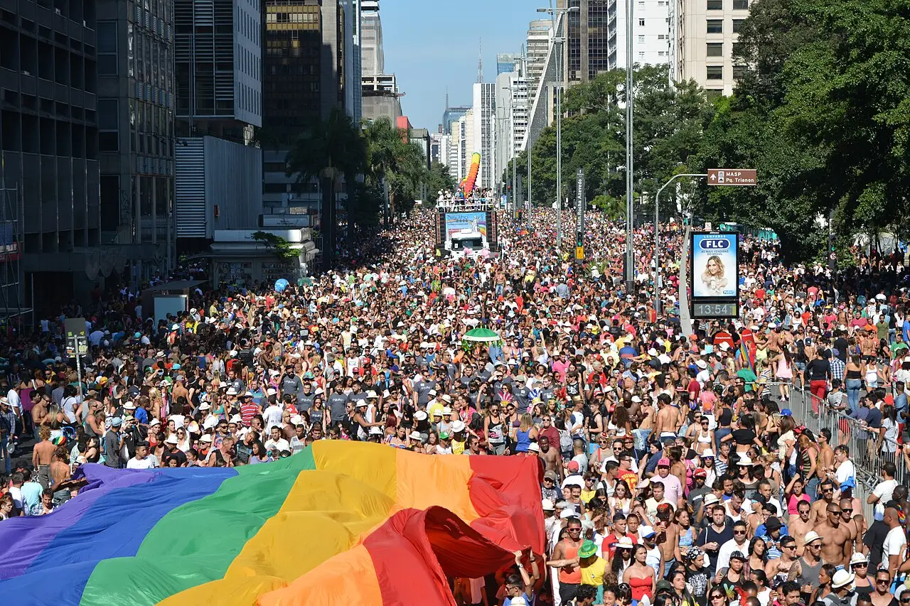 Uma bandeira arco-íris ocupa a frente e a avenida atrás está cheia de gente, entre prédios altos.