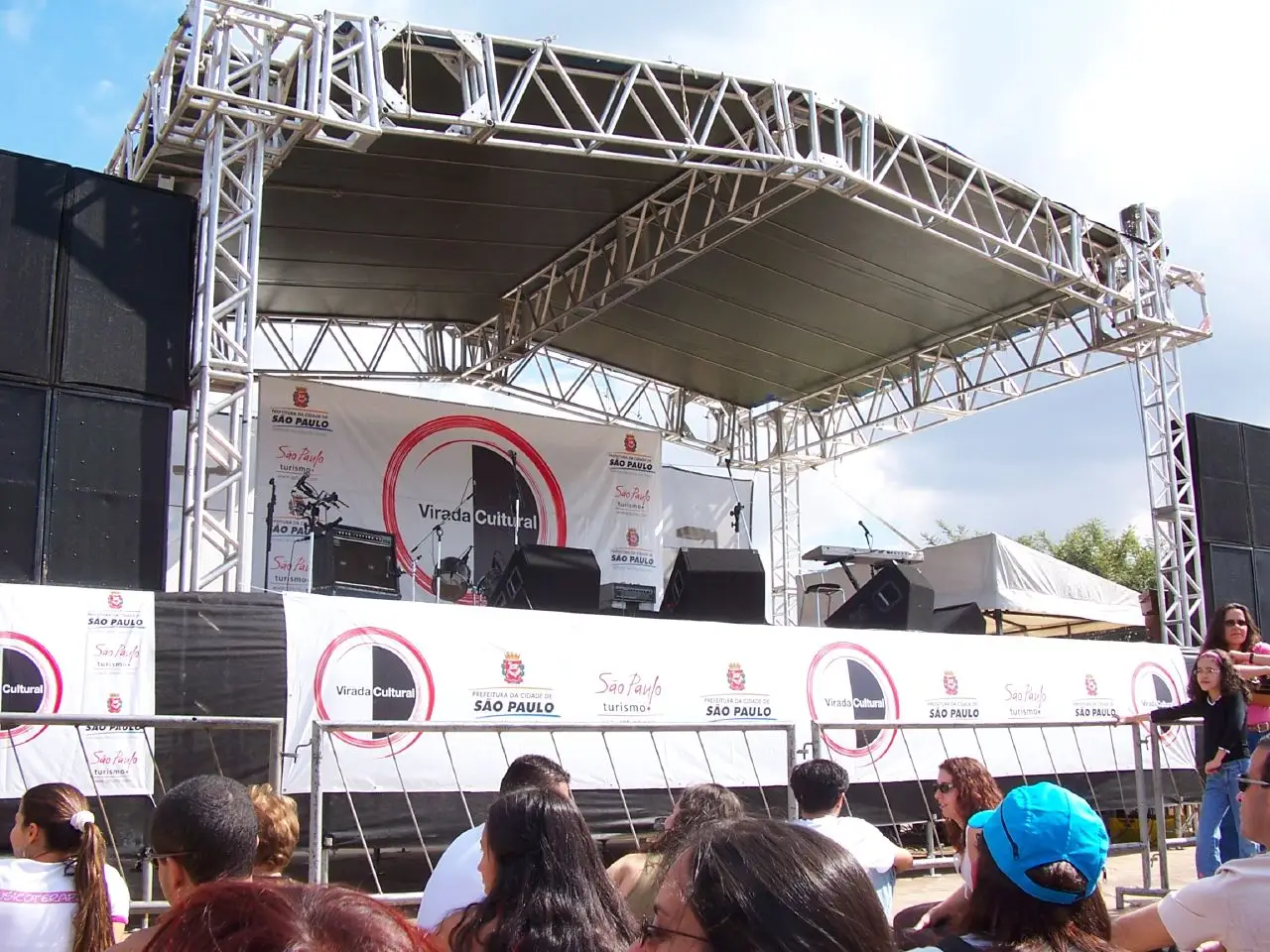 Pessoas olham um palco ao ar livre, e o painel atrás das caixas de som diz Virada Cultural.
