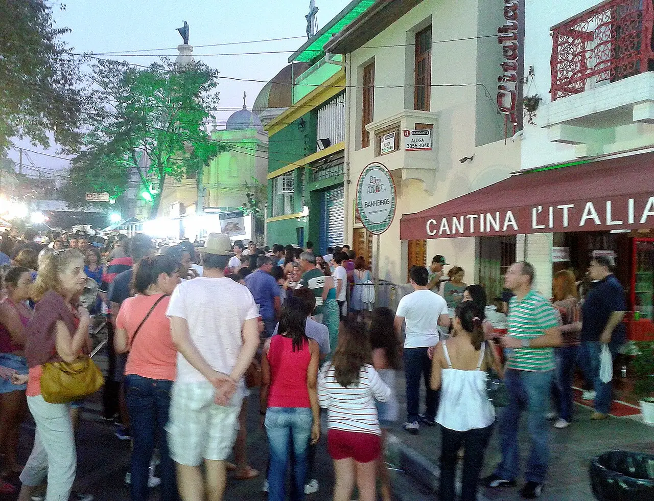 A multidão enche a rua ao anoitecer, diante da esquina com a placa Cantina Italia.