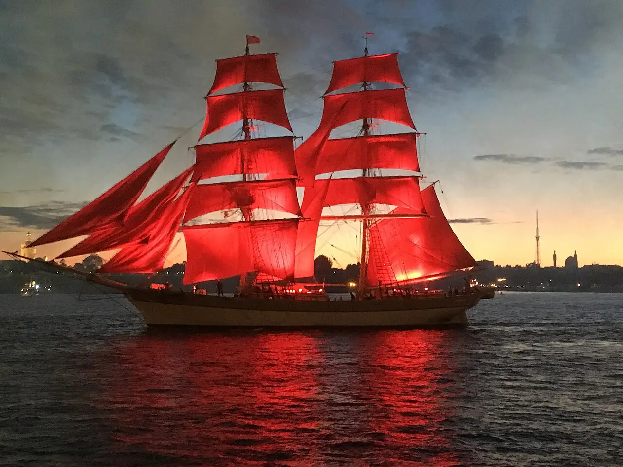 Um navio de velas vermelhas navega ao anoitecer, com o reflexo na água e uma torre no horizonte.