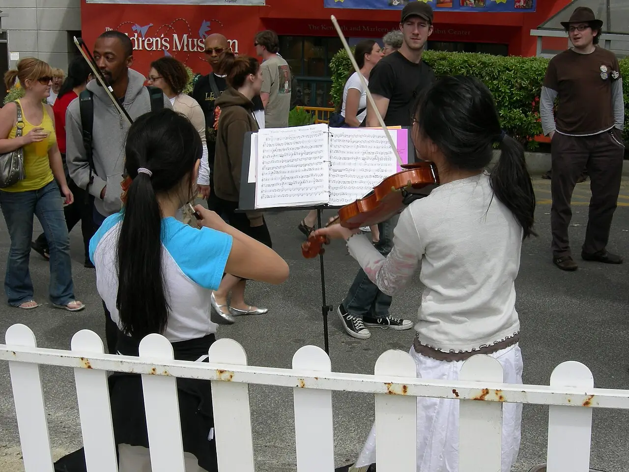 Deux violonistes jouent derrière une clôture blanche, avec des partitions sur les pupitres et des auditeurs autour.